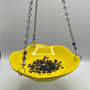 May include: A yellow ceramic bird feeder with a flower-shaped rim hangs from two silver chains with blue and white beads. The feeder is filled with birdseed.