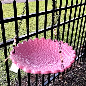 May include: A circular, pink, textured hanging bird bath with a scalloped edge. The basin features a detailed, embossed pattern across its surface and is suspended by three thin black metal chains that attach to the rim at evenly spaced intervals.