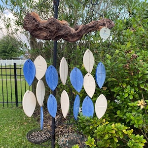 May include: A ceramic wind chime with blue and white leaf-shaped ornaments hanging from a driftwood branch. The chime is suspended from a black metal stand.