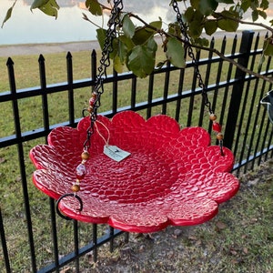 May include: Red ceramic bird feeder with a scalloped edge, hanging from a black chain with brown and red beads. The feeder is textured with a scaly pattern.