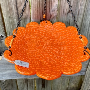 May include: Orange ceramic bird feeder with a scalloped edge and a crocodile skin pattern. The feeder hangs from a black chain with gold and brown beads.