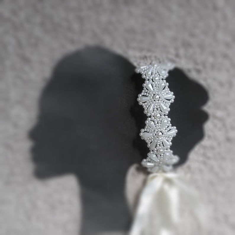 May include: A white beaded headband with a floral design. The headband is made of small white pearls and is arranged in a delicate floral pattern.