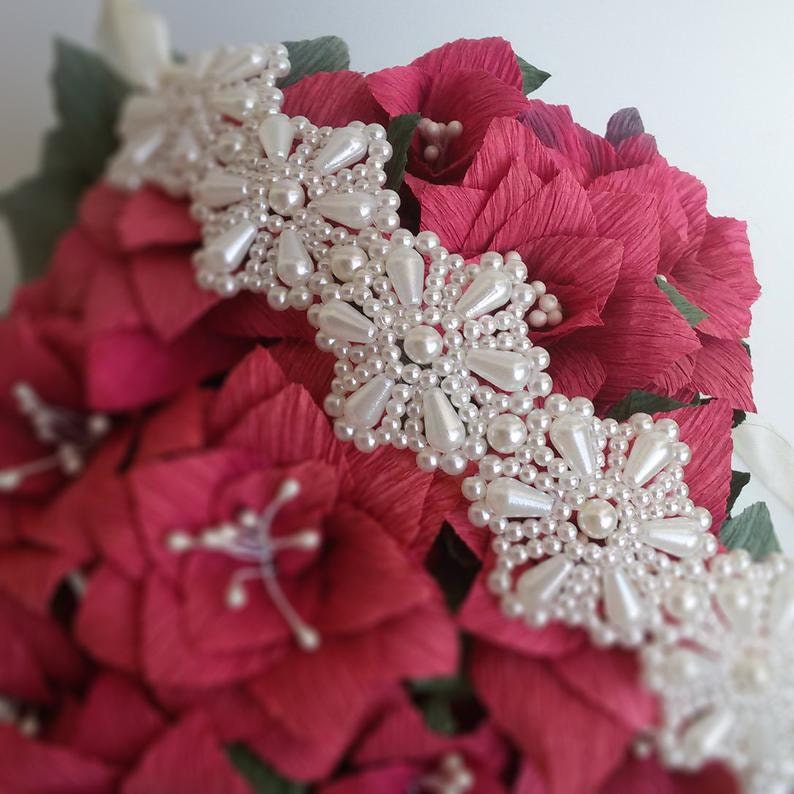 May include: A white beaded headband with a floral design, featuring multiple pearl-like beads arranged in a repeating pattern. The headband is displayed on a bouquet of red paper flowers.
