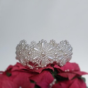 May include: A white beaded bracelet with a floral design. The bracelet is made of small white pearls and is arranged in a circular pattern. The bracelet is sitting on a red flower.