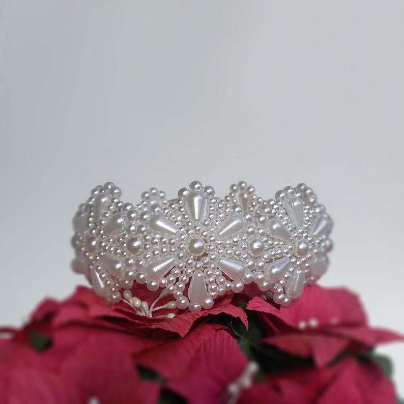 May include: A white beaded bracelet with a floral design. The bracelet is made of small white pearls and is arranged in a circular pattern. The bracelet is sitting on a red flower.