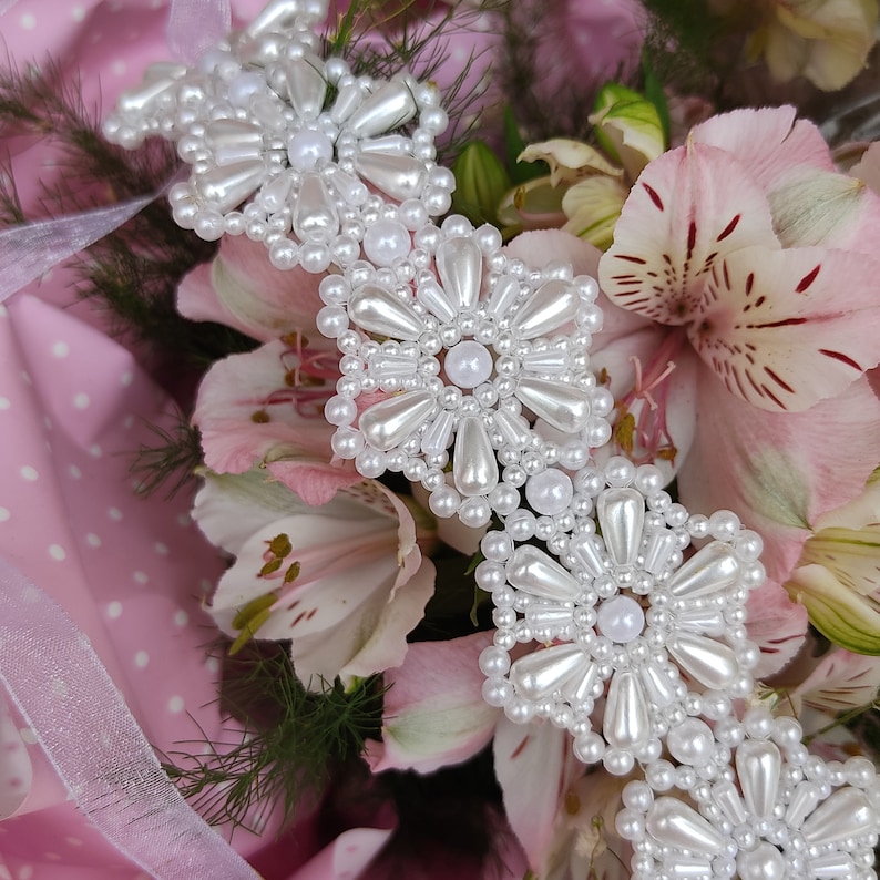 May include: A white pearl trim with a flower design. The trim is made of small white pearls and is arranged in a repeating pattern of flowers. The trim is shown against a pink background with white polka dots.