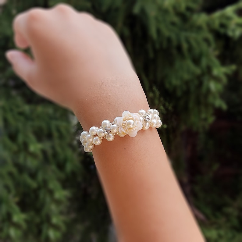 以下が含まれることがあります： 白い真珠のブレスレット、花のアクセント付き。ブレスレットは、小さな丸い真珠の複数のストランドが特徴です。真珠の中心と小さな透明なクリスタルが付いた白い花がブレスレットの中央にあります。ブレスレットは手首に着用されています。