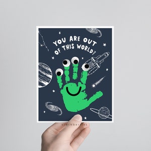 Pode incluir: Uma impressão de mão verde com olhos saltitantes e um rosto sorridente em um fundo azul escuro com estrelas brancas, planetas e um foguete. O texto "You are out of this world!" está escrito em letras brancas.