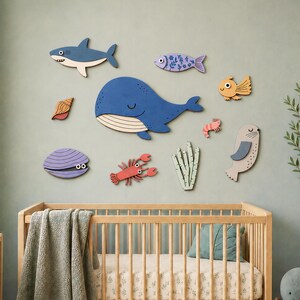 May include: A collection of wooden wall decals featuring a blue whale, a shark, a seal, a crab, a seashell, a clam, a small fish, and seaweed. The set includes the text kiwinberries EPS Ai SVG DXF, displayed above a wooden crib with a green blanket.
