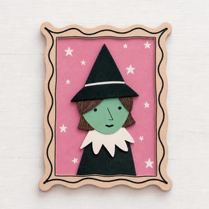 May include: A framed wall art piece featuring a green-faced witch with brown hair, a black pointed hat, and a white collar against a pink background with white stars. The frame has a wavy edge. Text at the bottom reads kiwinberries EPS Ai SVG DXF.