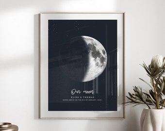 以下が含まれることがあります： 暗い青色の夜空を背景にした月の詳細なイラストが描かれた額入りのプリント。 「Our moon」というテキストと名前、日付が表示されています。 プリントは木製の表面に表示されています。