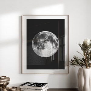 May include: A black and white photograph of a full moon in a black frame. The moon is the main focus of the image and is surrounded by a dark background.