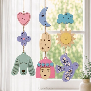 May include: Three vertical hanging decorations made of flat, painted wooden shapes suspended by twine. The shapes include a pink heart, a blue crescent moon, a light blue cloud, a purple flower, a hand, a yellow sun, a dog, a face, and a bird.