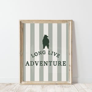 Pode incluir: Uma impressão emoldurada com um fundo listrado cinza e branco. Uma silhueta de urso preto fica no centro da impressão. O texto "Long Live Adventure" é impresso em verde abaixo do urso.