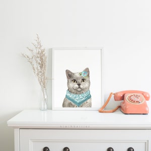 Puede incluir: Una pintura de acuarela de un gato gris con un pa&ntilde;uelo azul claro y una peque&ntilde;a flor en la cabeza. El gato est&aacute; mirando directamente al espectador. La pintura est&aacute; enmarcada en un marco blanco y est&aacute; colgada en una pared blanca.
