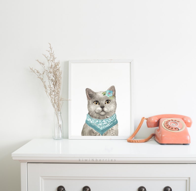 Puede incluir: Una pintura de acuarela de un gato gris con un pa&ntilde;uelo azul claro y una peque&ntilde;a flor en la cabeza. El gato est&aacute; mirando directamente al espectador. La pintura est&aacute; enmarcada en un marco blanco y est&aacute; colgada en una pared blanca.
