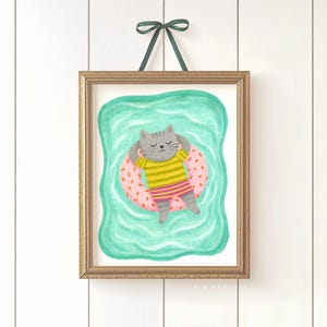 May include: A framed art print featuring a gray cat relaxing on a pink inflatable ring in a turquoise pool. The cat wears a yellow and green striped shirt. The artwork is framed in a gold-colored frame and is suspended by a green ribbon.