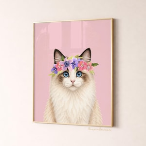 May include: A framed print featuring a Ragdoll cat with blue eyes and a floral crown. The cat has a white and tan coat, set against a pink background. The print is in a gold frame, with the artist's signature in the bottom right corner.