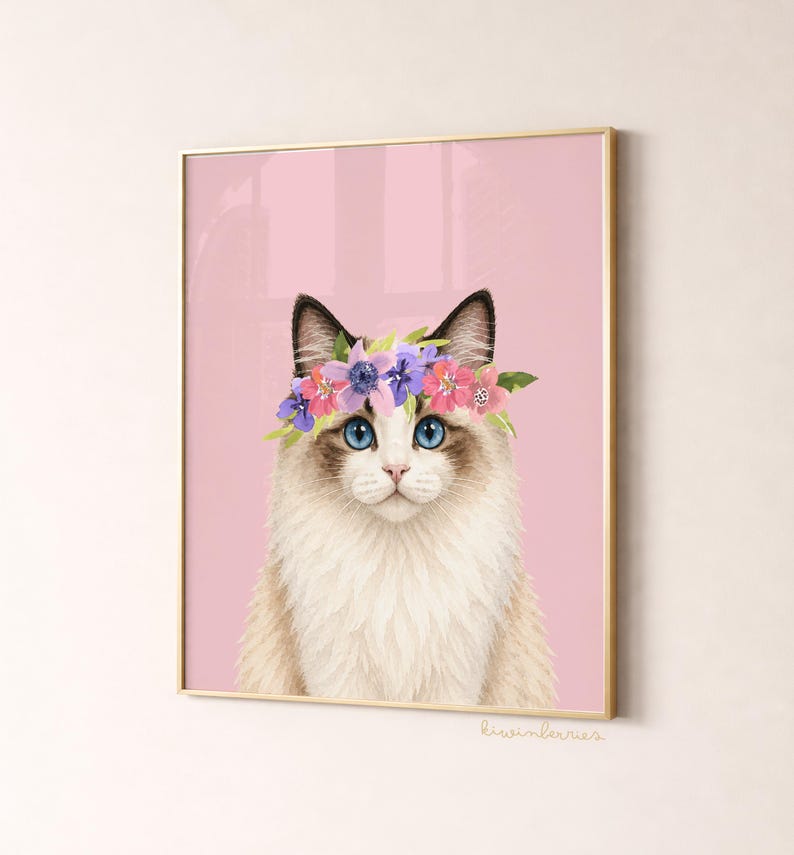 May include: A framed print featuring a Ragdoll cat with blue eyes and a floral crown. The cat has a white and tan coat, set against a pink background. The print is in a gold frame, with the artist's signature in the bottom right corner.