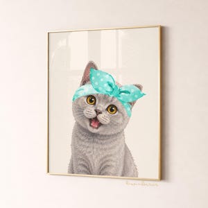 May include: A framed print featuring a gray cat with yellow eyes wearing a teal polka dot headband. The cat is smiling with its mouth open. The print has a gold frame and is set against a neutral background.