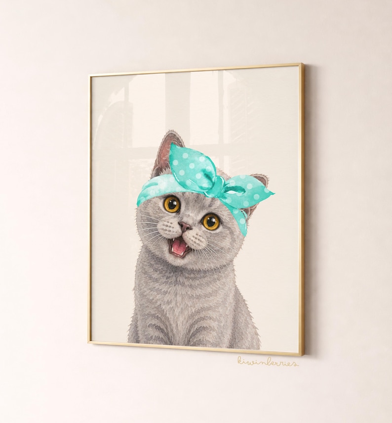 May include: A framed print featuring a gray cat with yellow eyes wearing a teal polka dot headband. The cat is smiling with its mouth open. The print has a gold frame and is set against a neutral background.