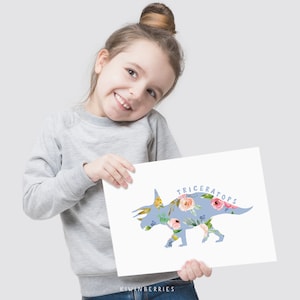 May include: A young girl holds up a blue and white print of a Triceratops dinosaur with floral accents. The print says "Triceratops" and "Kiwiberries" at the bottom.