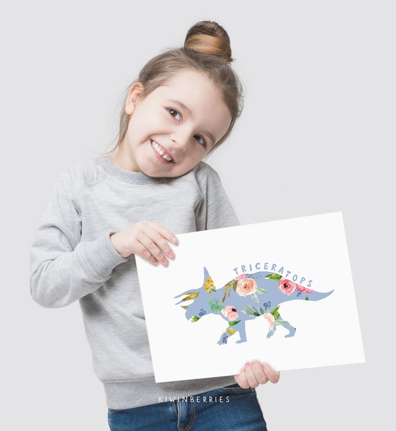 May include: A young girl holds up a blue and white print of a Triceratops dinosaur with floral accents. The print says "Triceratops" and "Kiwiberries" at the bottom.