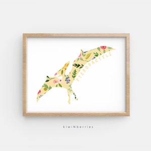 May include: A yellow floral print of a pterodactyl with the word "PTERODACTYL" written on the wing. The print is on a white background and is framed in a light wood frame.