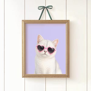 May include: A framed print featuring a white cat wearing pink heart-shaped sunglasses against a lavender background. The print is in a gold frame and is hanging from a green ribbon. The cat is the focal point of the artwork.