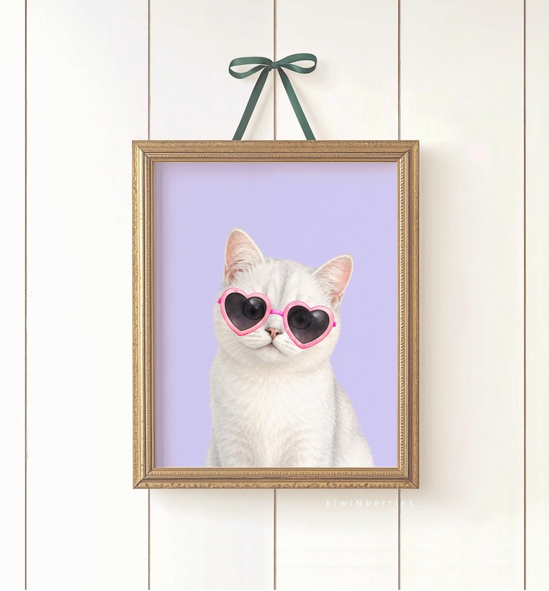 May include: A framed print featuring a white cat wearing pink heart-shaped sunglasses against a lavender background. The print is in a gold frame and is hanging from a green ribbon. The cat is the focal point of the artwork.