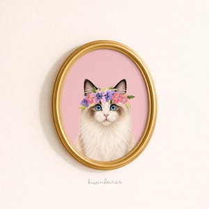 May include: An oval framed print featuring a Ragdoll cat with blue eyes and a floral crown. The cat is set against a pink background, within a gold-colored frame. The artist's signature, "kiwinberries," is at the bottom.