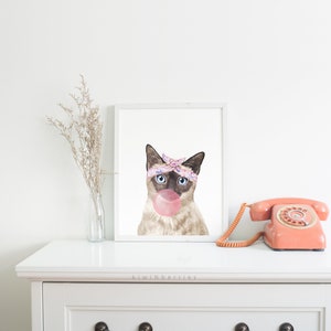 Puede incluir: Una ilustraci&oacute;n de acuarela de un gato siam&eacute;s con un pa&ntilde;uelo rosa y morado y soplando una burbuja rosa. El gato tiene ojos azules y mira directamente al espectador. El fondo es blanco.