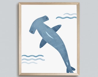 May include: A blue hammerhead shark illustration with a white background and blue wavy lines representing water. The illustration is framed in a light wood frame. The text "kiwiNberries" is at the bottom of the image.