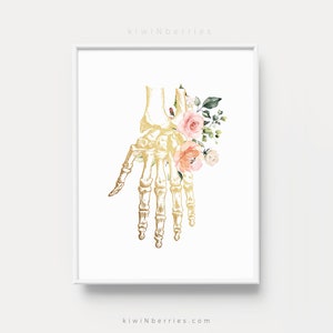 Puede incluir: Un esqueleto de mano anat&oacute;mico dorado con flores de acuarela rosa y melocot&oacute;n. Las flores est&aacute;n dispuestas alrededor de los huesos de la mano y la mu&ntilde;eca. El fondo es blanco.