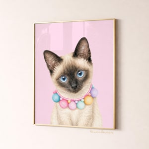 May include: A framed print featuring a Siamese cat with striking blue eyes and a colorful pom-pom collar. The cat is set against a soft pink background, creating a whimsical and charming aesthetic. The artwork is signed in the bottom right corner.