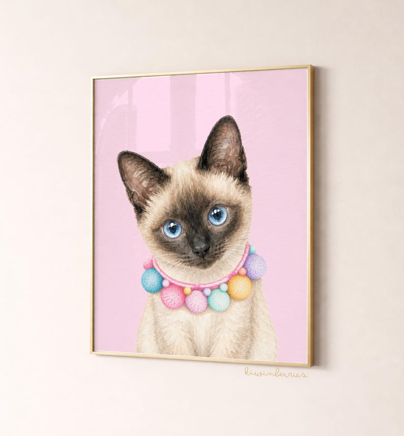 May include: A framed print featuring a Siamese cat with striking blue eyes and a colorful pom-pom collar. The cat is set against a soft pink background, creating a whimsical and charming aesthetic. The artwork is signed in the bottom right corner.