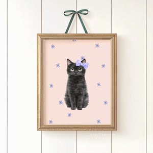 May include: A framed art print featuring a black cat with a purple bow, set against a pink background with blue snowflake accents. The print is in a gold frame and is suspended by a green ribbon.