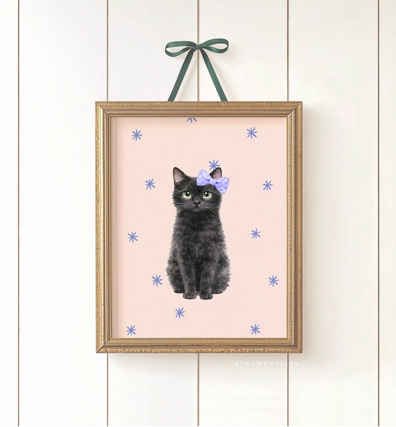 May include: A framed art print featuring a black cat with a purple bow, set against a pink background with blue snowflake accents. The print is in a gold frame and is suspended by a green ribbon.