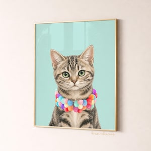 May include: A framed print featuring a tabby cat with green eyes and a colorful pom-pom collar. The cat is set against a light teal background. The frame is gold. The print is signed "liwiberries".