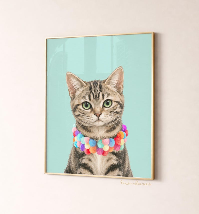 May include: A framed print featuring a tabby cat with green eyes and a colorful pom-pom collar. The cat is set against a light teal background. The frame is gold. The print is signed "liwiberries".