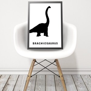 Op de afbeelding: Zwart-witte silhouet van een Brachiosaurus-dinosaurus op een witte achtergrond. De naam van de dinosaurus is in zwarte tekst onder de silhouet gedrukt. De afbeelding is ingelijst en wordt op een witte stoel getoond.