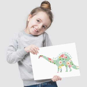 May include: A young girl holds up a floral print dinosaur art print. The print features a green brachiosaurus silhouette with a floral pattern. The text "Brachiosaurus" is written in green cursive on the print.