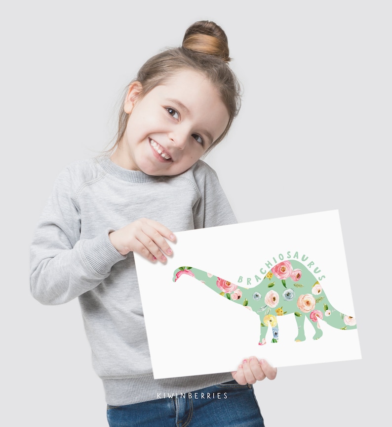 May include: A young girl holds up a floral print dinosaur art print. The print features a green brachiosaurus silhouette with a floral pattern. The text "Brachiosaurus" is written in green cursive on the print.