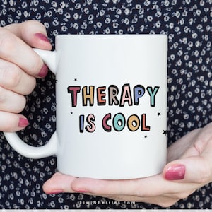May include: White ceramic mug with a colorful graphic that says "Therapy is Cool" in a playful font. The mug is held by a person's hands.