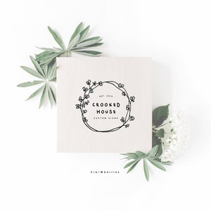 Op de afbeelding: Een witte kaart met een zwart-wit logo met een krans van bloemen en de tekst "EST. 2015 CROOKED HOUSE CUSTOM SIGNS" en de tekst "kiwiNberries" onder de kaart.