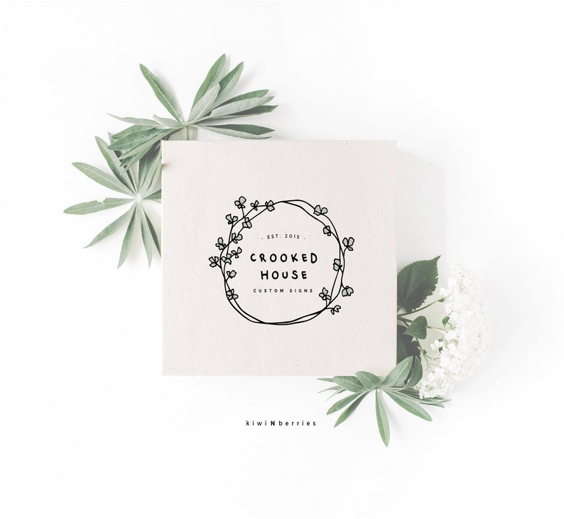 Op de afbeelding: Een witte kaart met een zwart-wit logo met een krans van bloemen en de tekst "EST. 2015 CROOKED HOUSE CUSTOM SIGNS" en de tekst "kiwiNberries" onder de kaart.