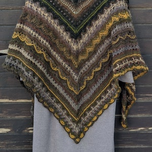 Puede incluir: Un chal de ganchillo triangular en tonos marrones, verdes y amarillos. El chal está colgado de una percha de madera sobre un fondo de madera oscura.