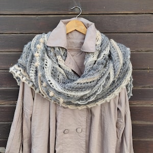 Könnte beinhalten: Ein grauer und weißer Häkel-Schal mit Fransenbordüre, über eine hellbraune Jacke gelegt.