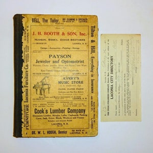 May include: An antique directory and a small receipt are displayed on a white surface. The directory has a yellowed cover with black text and advertisements for local businesses. The receipt has handwritten text.