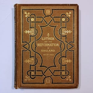 May include: A vintage book titled "Luther and the Reformation in England" with a brown cover. The cover features ornate gold and black decorative elements, including floral designs and geometric patterns. The book's spine is also brown.